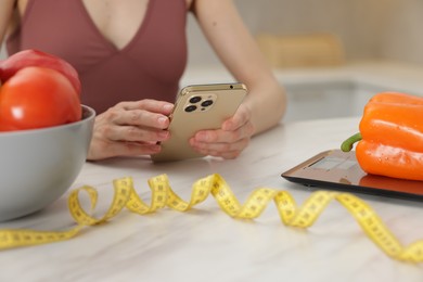 Photo of Woman using smartphone at white marble table with vegetables, kitchen scale and measuring tape indoors, closeup