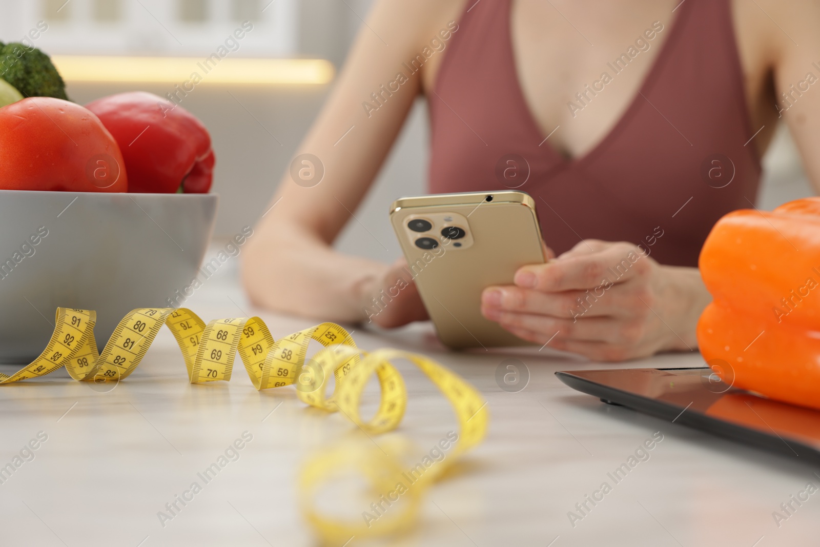 Woman using smartphone at white marble table with vegetables, kitchen scale and measuring tape indoors, closeup Photo of Woman using smartphone at white marble table with vegetables, kitchen scale and measuring tape indoors, closeup