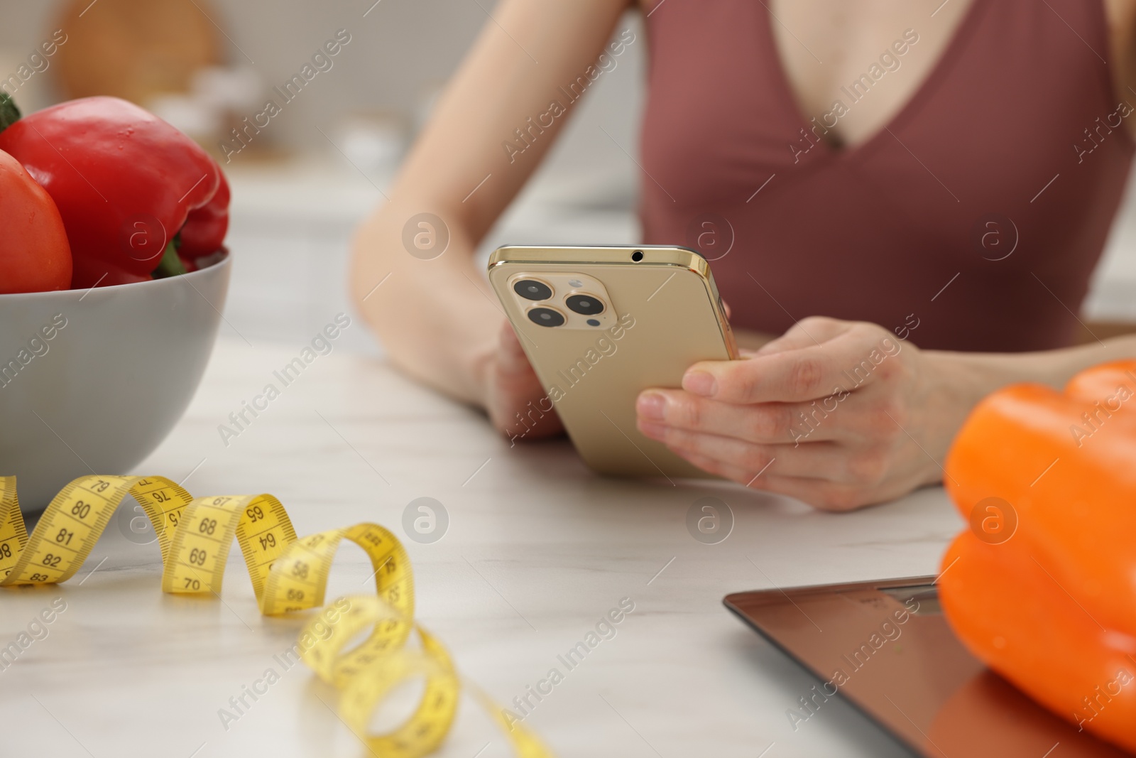Woman using smartphone at white marble table with vegetables, kitchen scale and measuring tape indoors, closeup Photo of Woman using smartphone at white marble table with vegetables, kitchen scale and measuring tape indoors, closeup