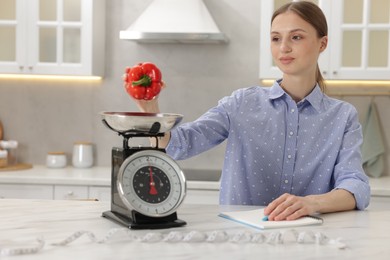 Photo of Woman weighting bell pepper on kitchen scale at white marble table indoors
