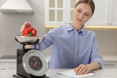 Photo of Woman weighting bell pepper on kitchen scale at white marble table indoors