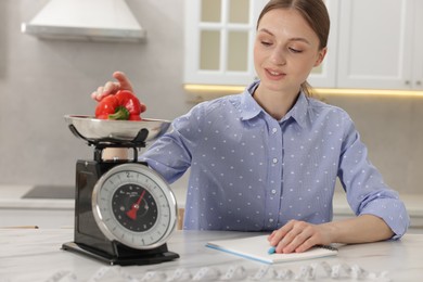 Photo of Woman weighting bell pepper on kitchen scale at white marble table indoors
