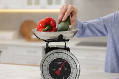 Photo of Woman weighting vegetables on kitchen scale at white table indoors, closeup