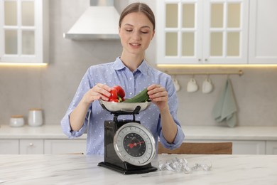Photo of Woman weighting vegetables on kitchen scale at white marble table indoors