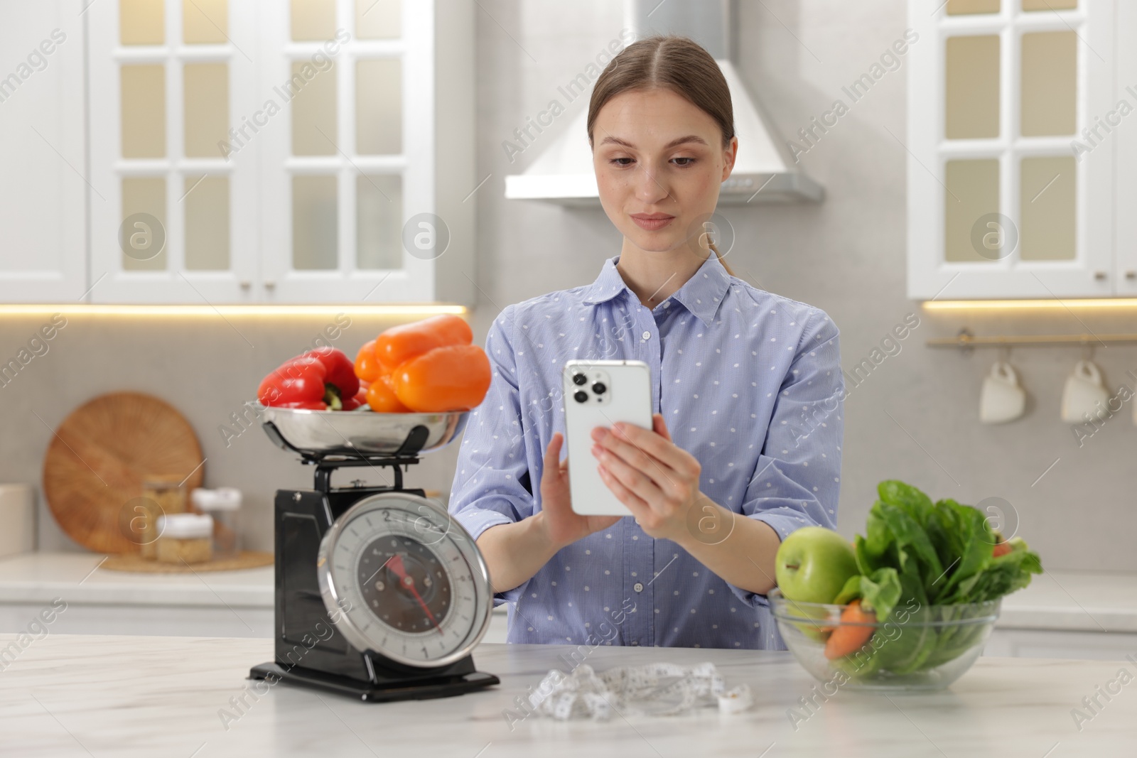 Woman using smartphone near white marble table with kitchen scale, healthy food and measuring tape indoors Photo of Woman using smartphone near white marble table with kitchen scale, healthy food and measuring tape indoors