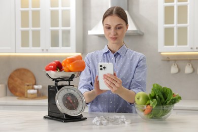 Photo of Woman using smartphone near white marble table with kitchen scale, healthy food and measuring tape indoors
