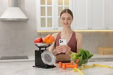 Photo of Woman using smartphone near white marble table with kitchen scale, healthy food, dumbbells and measuring tape indoors