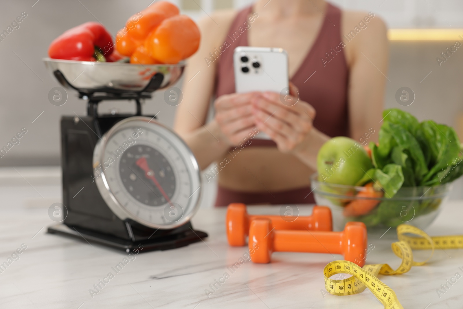 Woman using smartphone near white marble table with kitchen scale, healthy food, dumbbells and measuring tape indoors, selective focus Photo of Woman using smartphone near white marble table with kitchen scale, healthy food, dumbbells and measuring tape indoors, selective focus