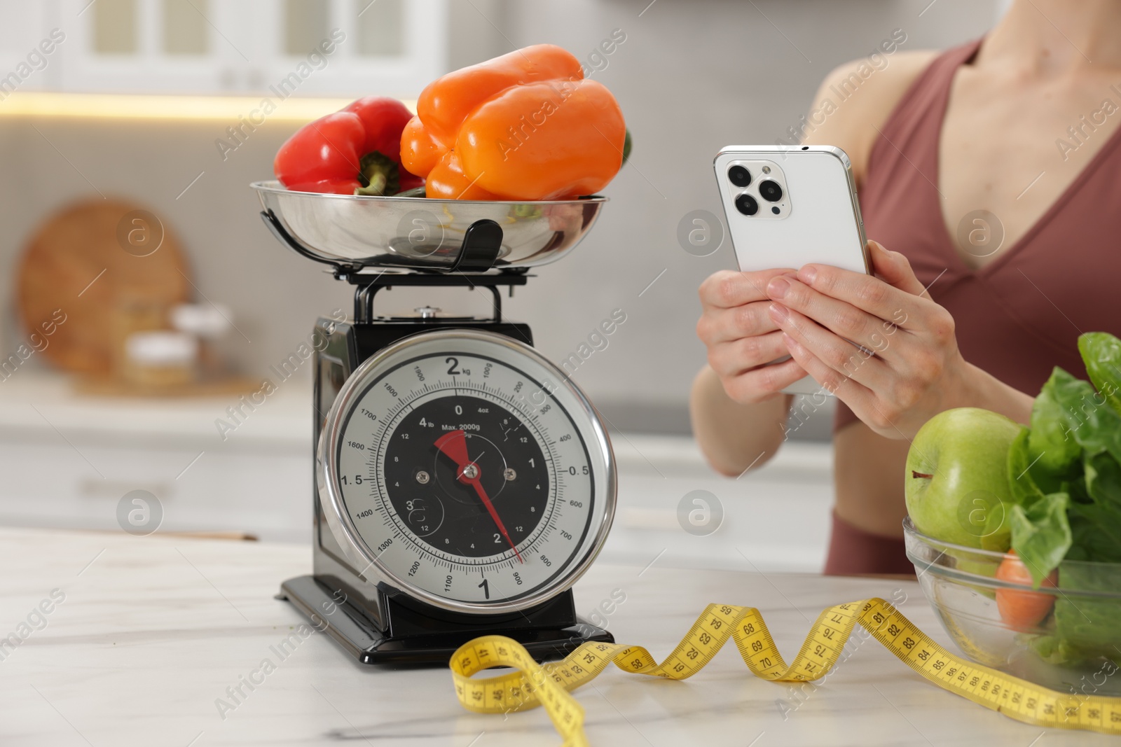 Woman using smartphone near white marble table with kitchen scale, healthy food and measuring tape indoors, closeup Photo of Woman using smartphone near white marble table with kitchen scale, healthy food and measuring tape indoors, closeup
