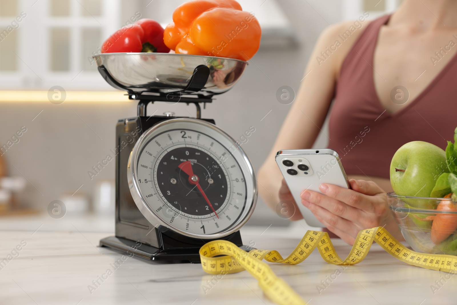 Woman using smartphone near white marble table with kitchen scale, healthy food and measuring tape indoors, closeup Photo of Woman using smartphone near white marble table with kitchen scale, healthy food and measuring tape indoors, closeup