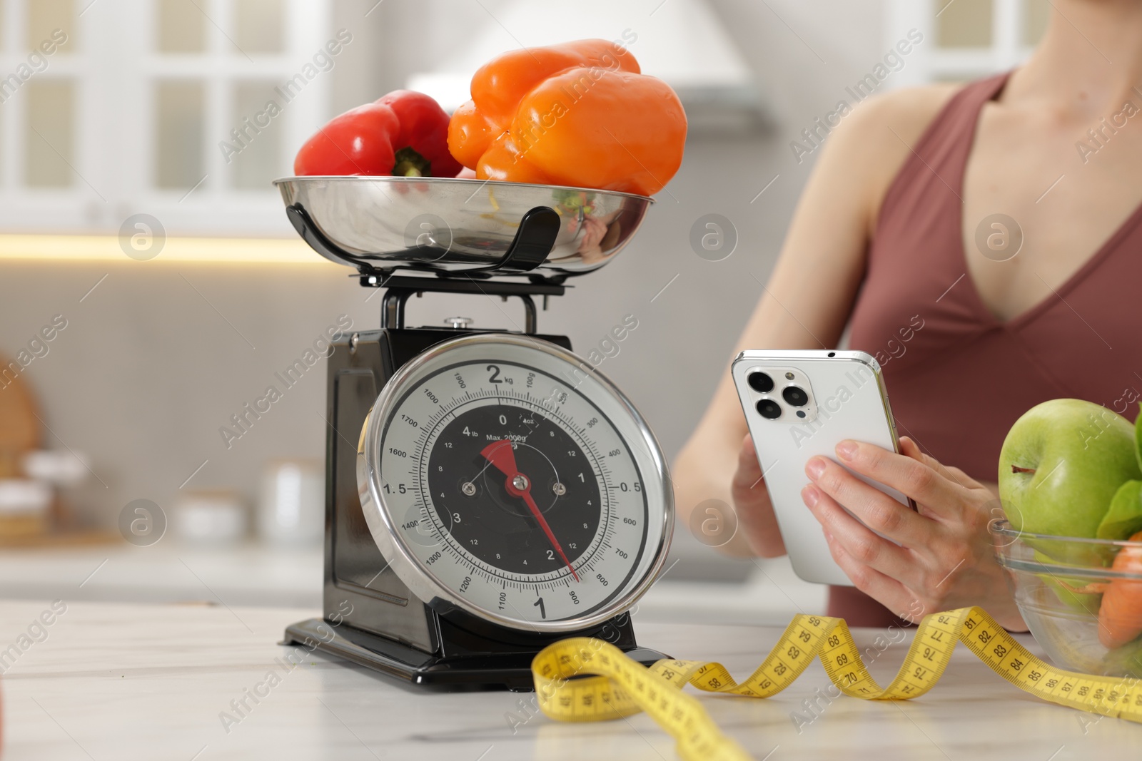 Woman using smartphone near white marble table with kitchen scale, healthy food and measuring tape indoors, closeup Photo of Woman using smartphone near white marble table with kitchen scale, healthy food and measuring tape indoors, closeup