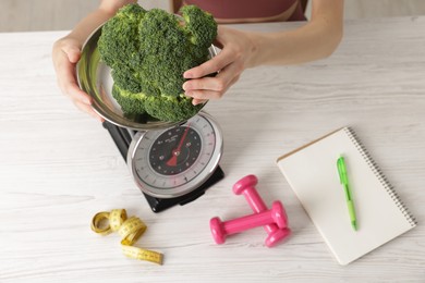 Photo of Woman weighting broccoli on kitchen scale at light wooden table indoors, top view