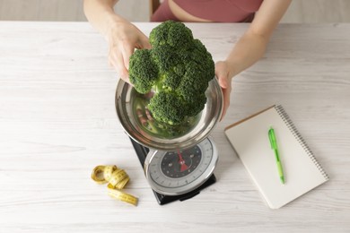 Photo of Woman weighting broccoli on kitchen scale at light wooden table indoors, top view