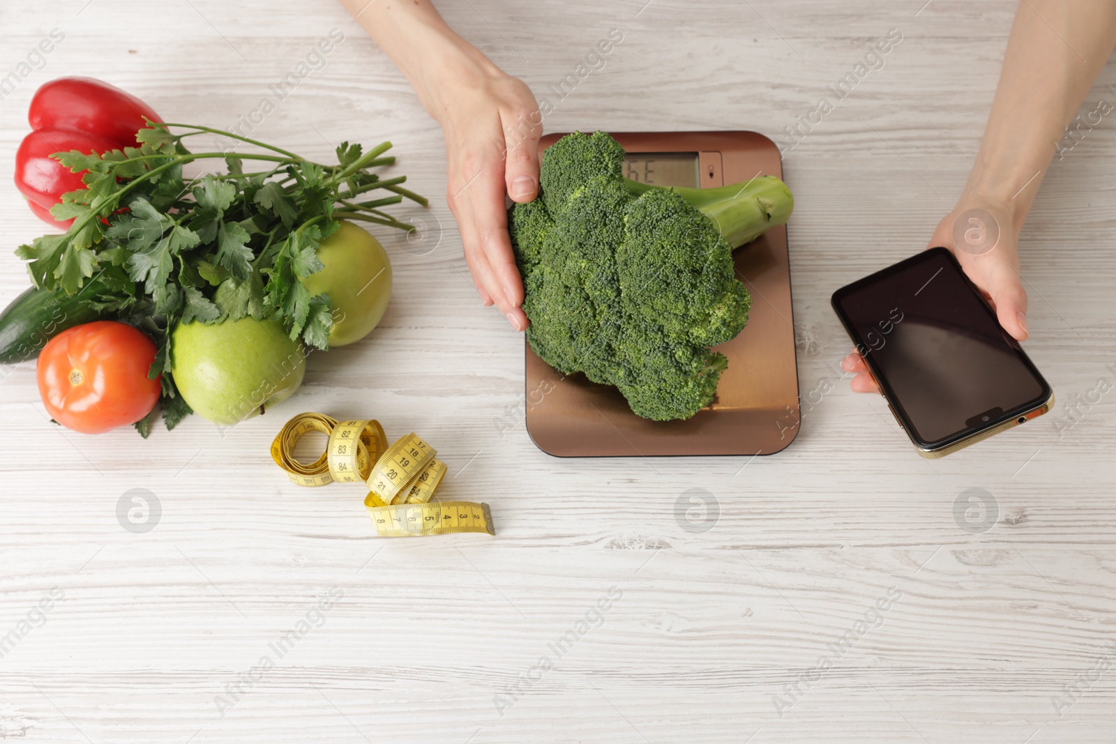 Woman with smartphone weighting broccoli on kitchen scale at light wooden table, top view Photo of Woman with smartphone weighting broccoli on kitchen scale at light wooden table, top view