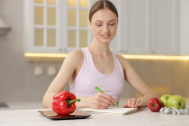 Photo of Woman developing diet plan at white wooden table indoors, focus on kitchen scale and bell pepper