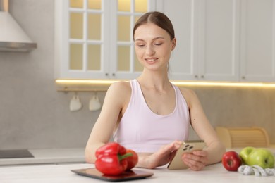 Photo of Woman using smartphone at white table with kitchen scale, healthy food and measuring tape indoors