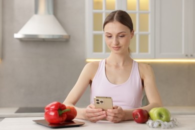 Photo of Woman using smartphone at white table with kitchen scale, healthy food and measuring tape indoors