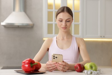 Photo of Woman using smartphone at white table with kitchen scale, healthy food and measuring tape indoors