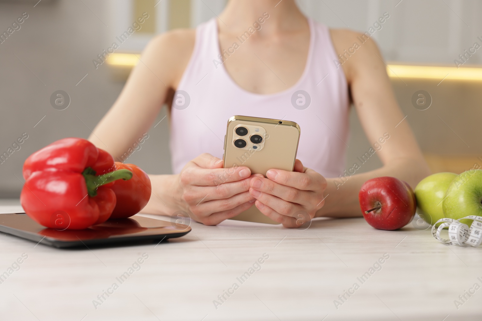 Woman using smartphone at white wooden table with kitchen scale, healthy food and measuring tape indoors, closeup Photo of Woman using smartphone at white wooden table with kitchen scale, healthy food and measuring tape indoors, closeup