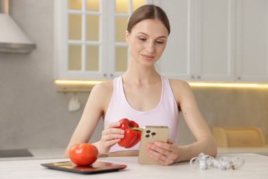Photo of Woman with smartphone and bell pepper at white table indoors