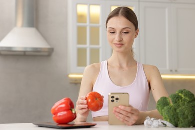 Photo of Woman with smartphone and tomato at white table indoors