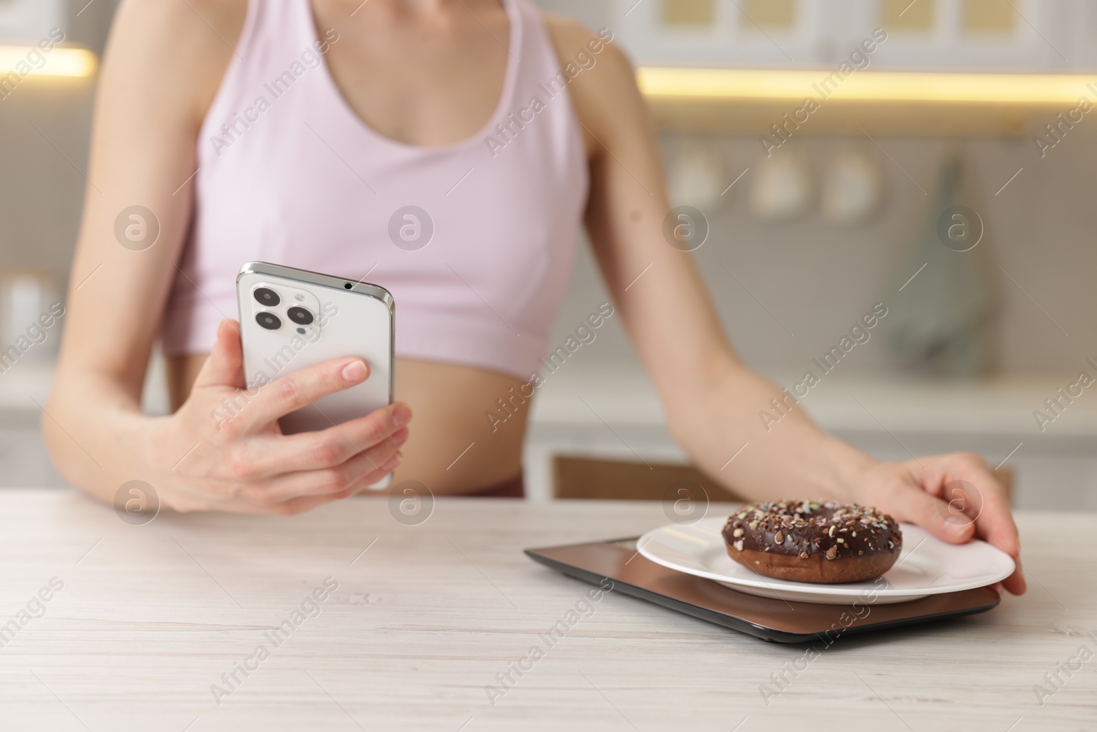 Woman with smartphone weighting donut on kitchen scale at white wooden table indoors, closeup Photo of Woman with smartphone weighting donut on kitchen scale at white wooden table indoors, closeup
