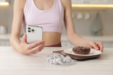 Photo of Woman with smartphone weighting donut on kitchen scale at white wooden table indoors, closeup