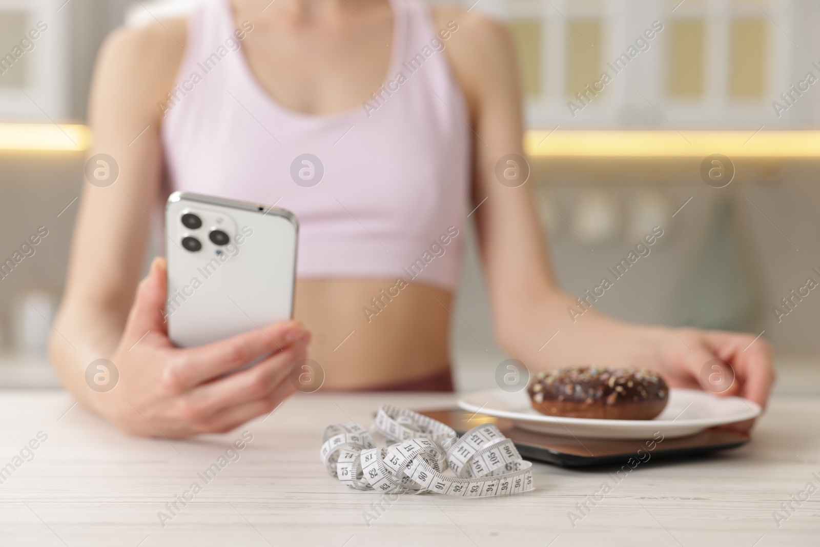 Woman with smartphone weighting donut on kitchen scale at white wooden table indoors, closeup Photo of Woman with smartphone weighting donut on kitchen scale at white wooden table indoors, closeup