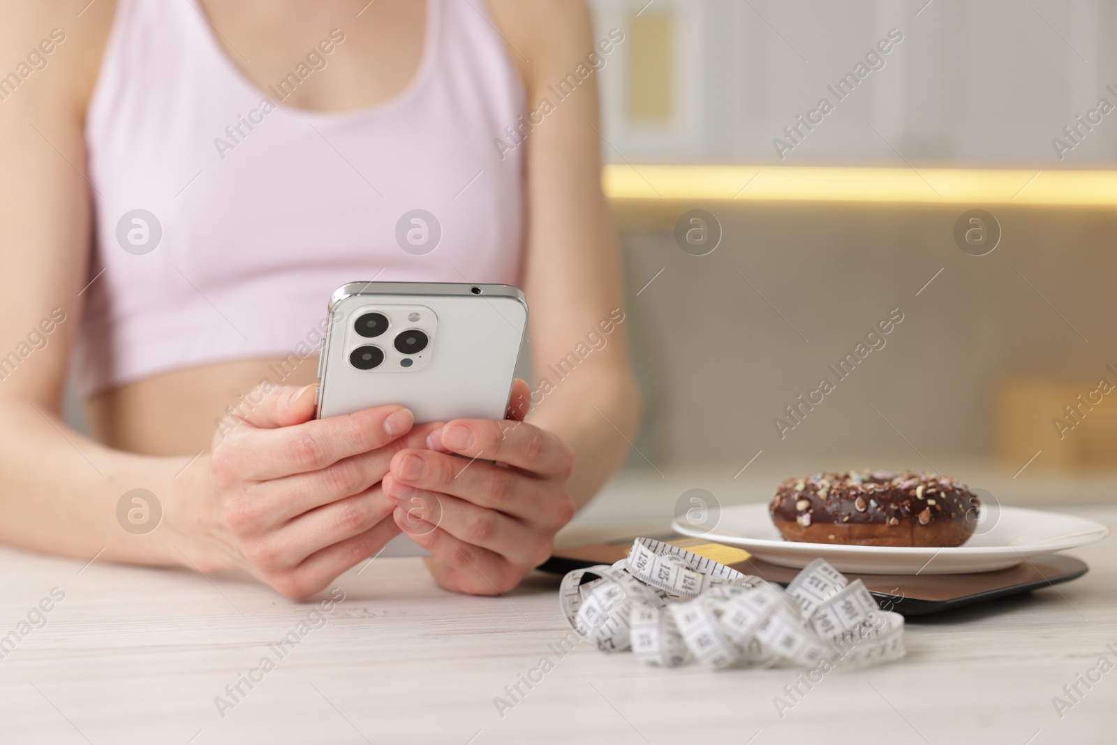 Woman using smartphone at white wooden table with kitchen scale, donut and measuring tape indoors, closeup Photo of Woman using smartphone at white wooden table with kitchen scale, donut and measuring tape indoors, closeup