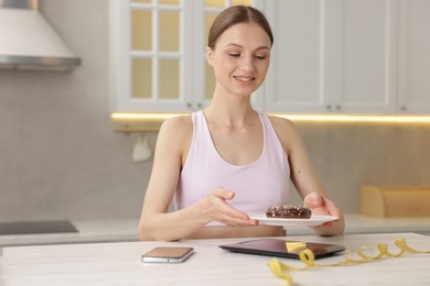 Photo of Woman weighting donut on kitchen scale at white table indoors