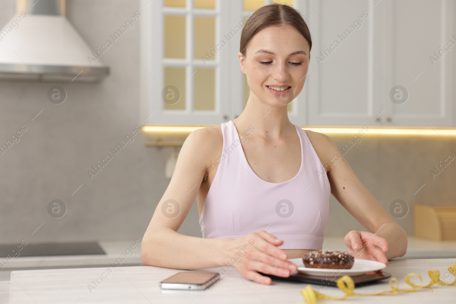 Photo of Woman weighting donut on kitchen scale at white table indoors, space for text