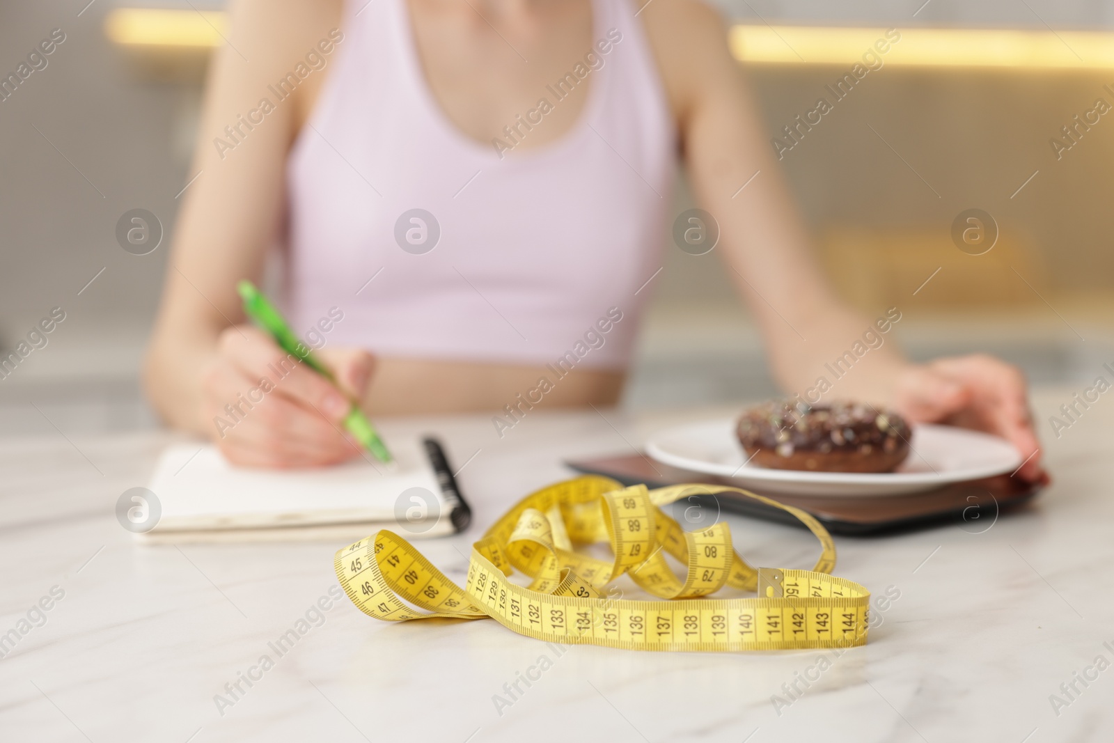 Woman developing diet plan while weighing donut on kitchen scale at white marble table indoors, selective focus Photo of Woman developing diet plan while weighing donut on kitchen scale at white marble table indoors, selective focus