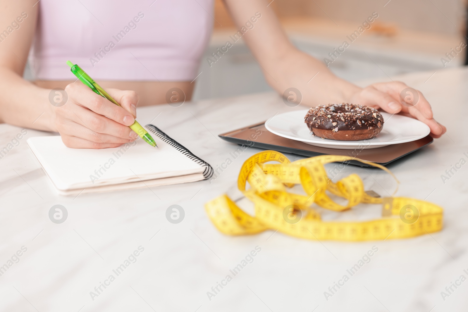 Woman developing diet plan while weighing donut on kitchen scale at white marble table indoors, closeup Photo of Woman developing diet plan while weighing donut on kitchen scale at white marble table indoors, closeup