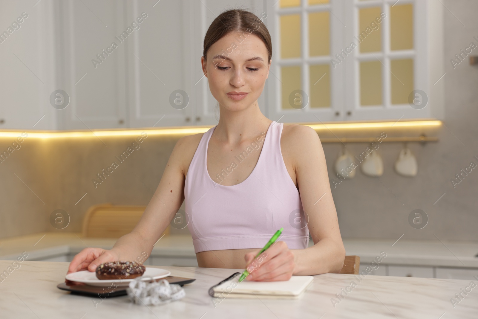 Woman developing diet plan while weighing donut on kitchen scale at white marble table indoors Photo of Woman developing diet plan while weighing donut on kitchen scale at white marble table indoors