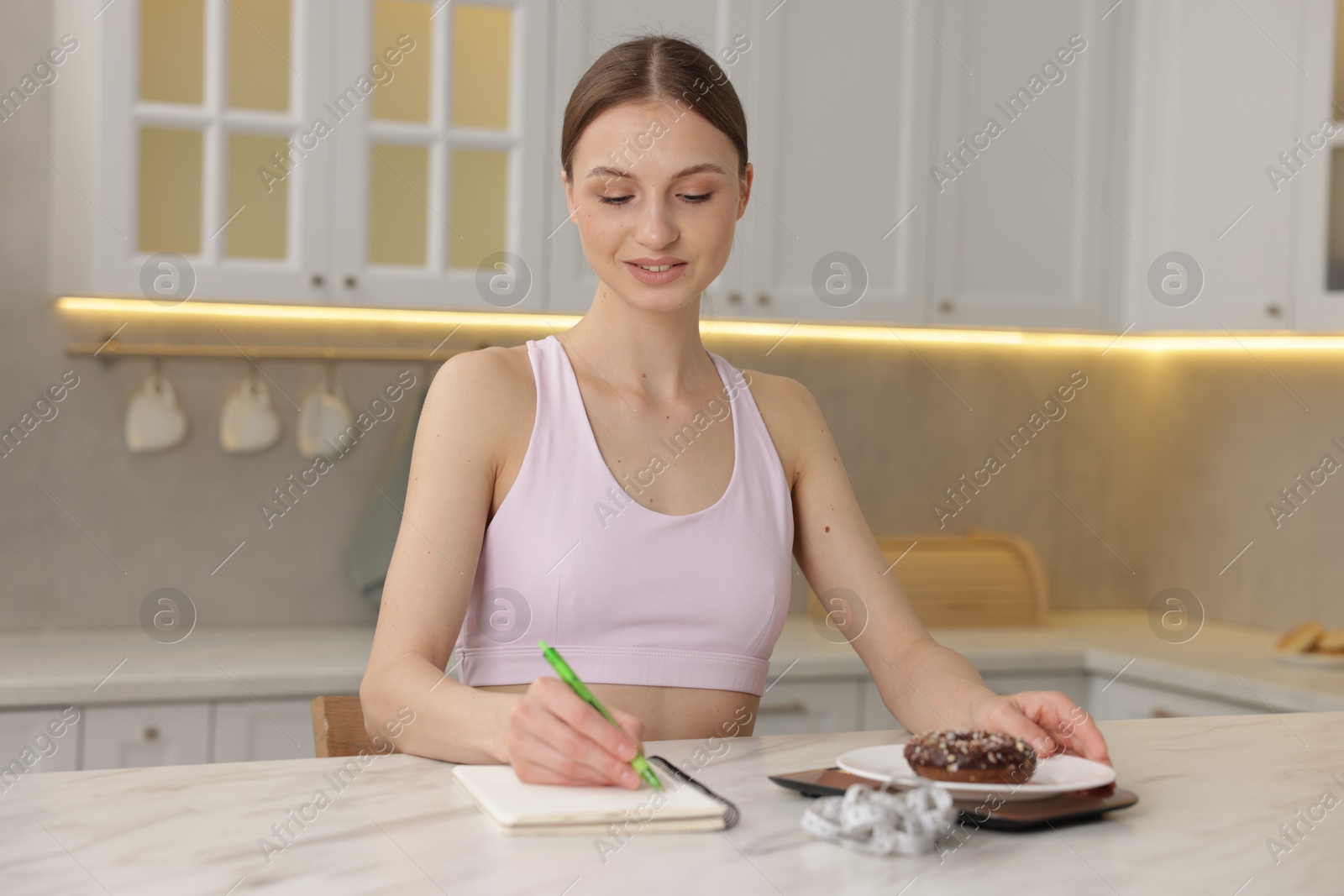Woman developing diet plan while weighing donut on kitchen scale at white marble table indoors Photo of Woman developing diet plan while weighing donut on kitchen scale at white marble table indoors