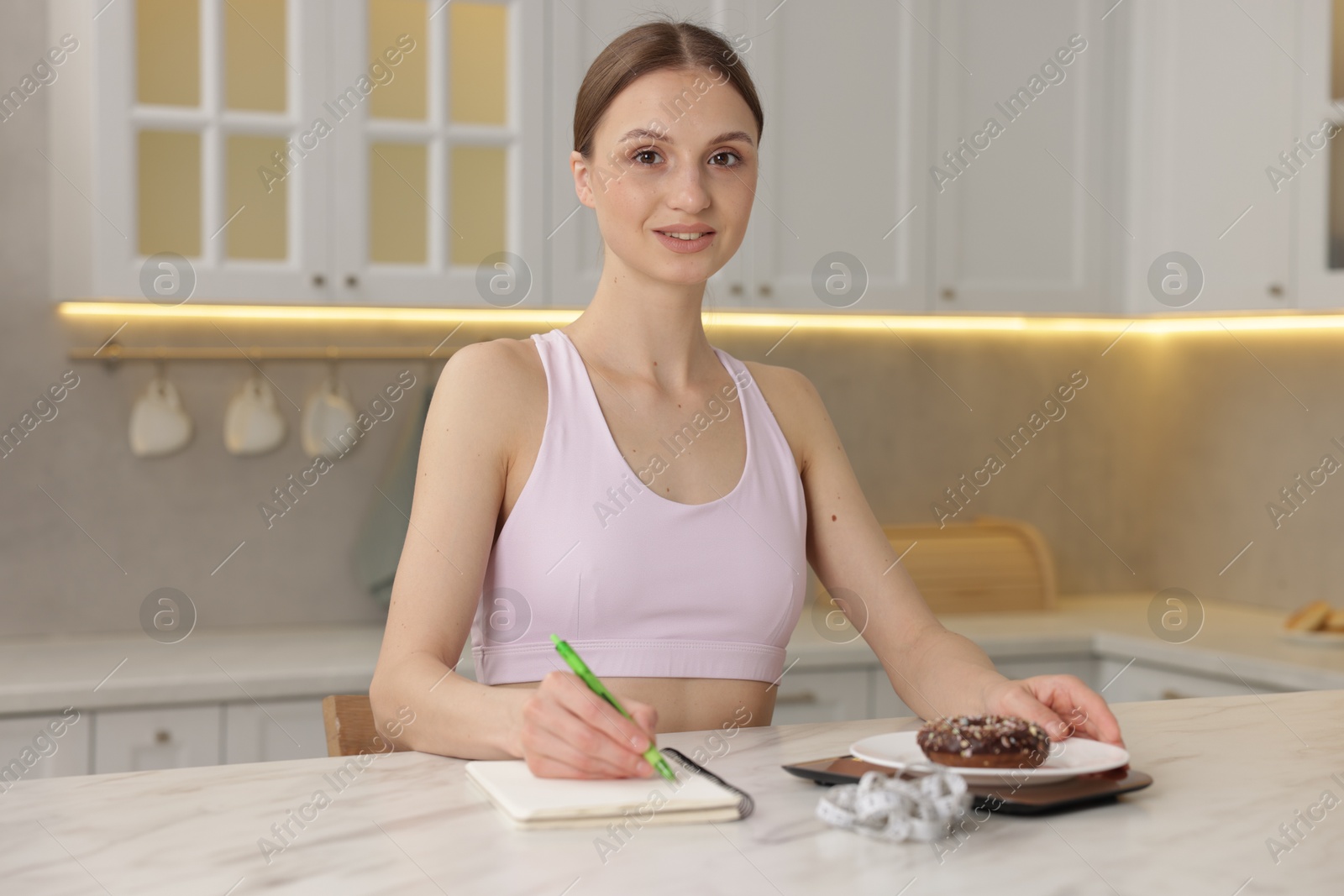Woman developing diet plan while weighing donut on kitchen scale at white marble table indoors Photo of Woman developing diet plan while weighing donut on kitchen scale at white marble table indoors