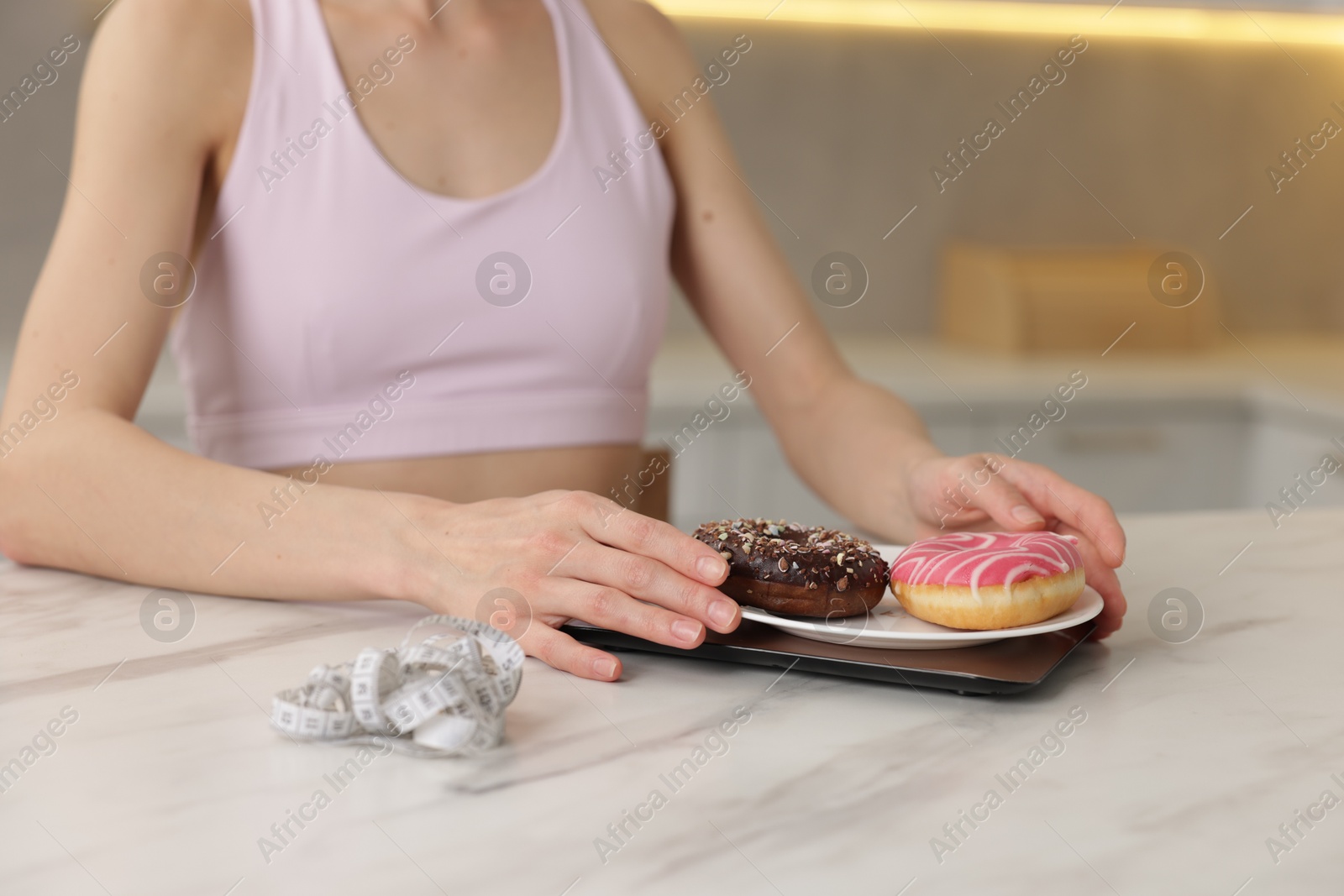 Photo of Woman weighing donuts on kitchen scale at white marble table indoors, closeup