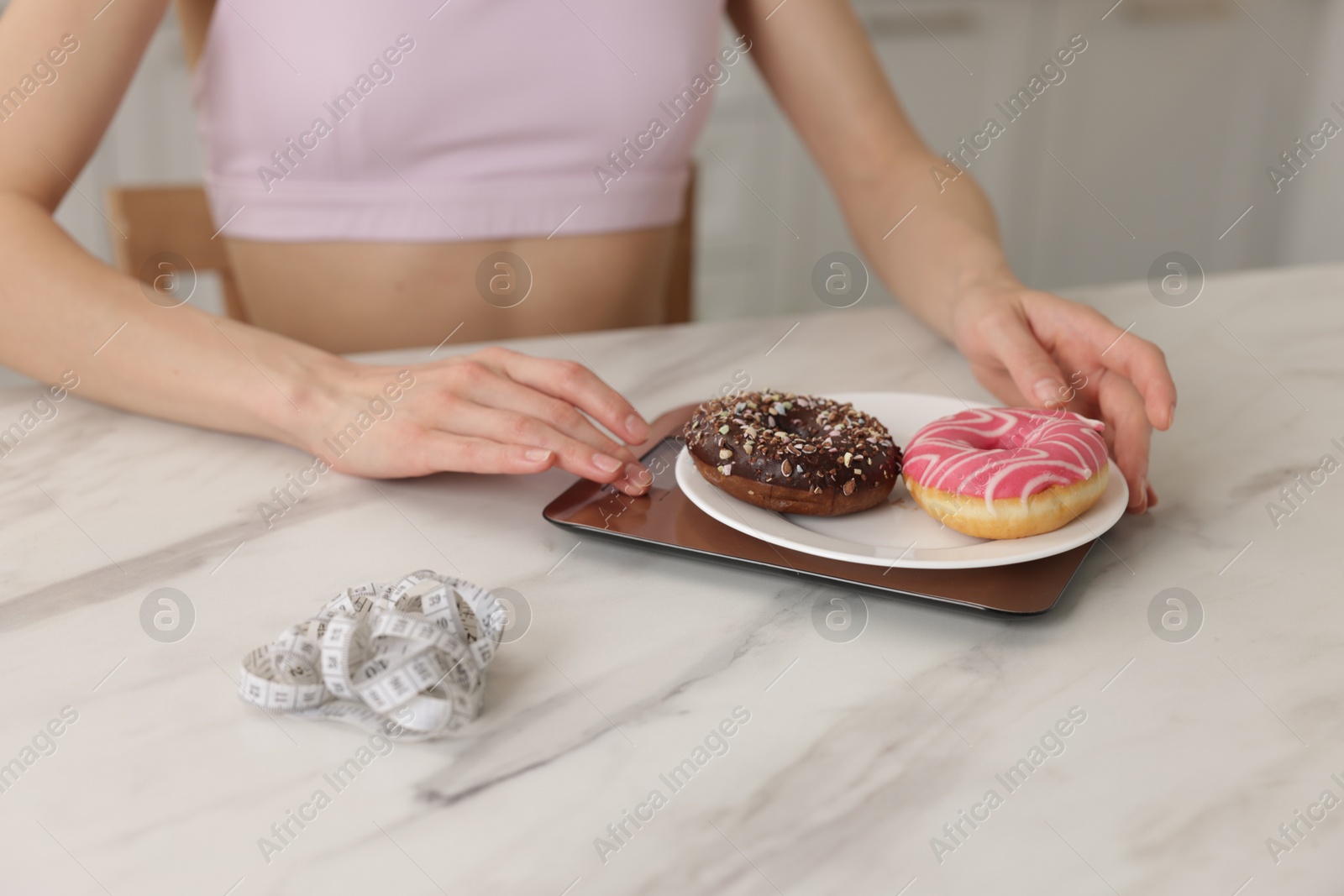 Photo of Woman weighing donuts on kitchen scale at white marble table indoors, closeup