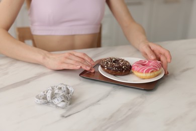Photo of Woman weighing donuts on kitchen scale at white marble table indoors, closeup