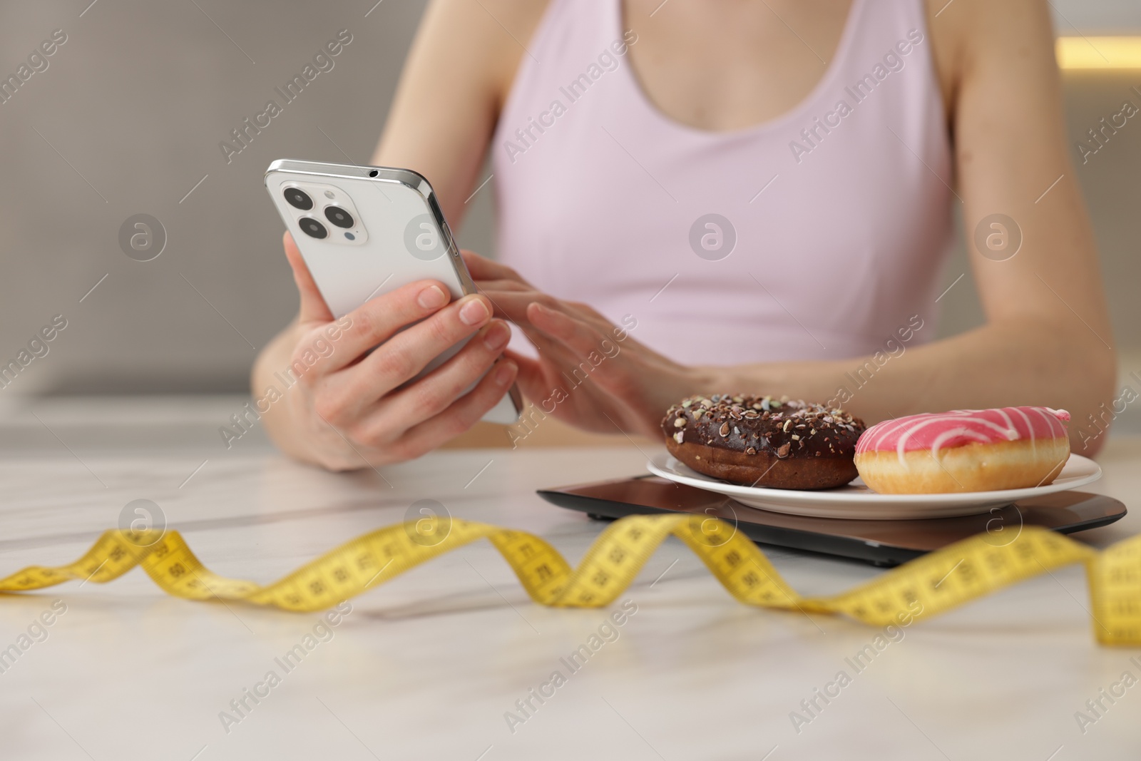 Photo of Woman using smartphone at white marble table with kitchen scale, donuts and measuring tape indoors, closeup