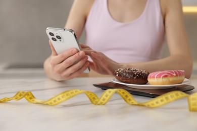 Photo of Woman using smartphone at white marble table with kitchen scale, donuts and measuring tape indoors, closeup