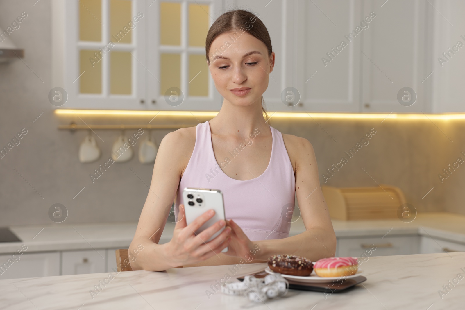 Woman using smartphone at white marble table with kitchen scale, donuts and measuring tape indoors Photo of Woman using smartphone at white marble table with kitchen scale, donuts and measuring tape indoors