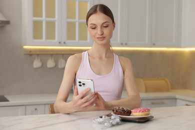 Photo of Woman using smartphone at white marble table with kitchen scale, donuts and measuring tape indoors