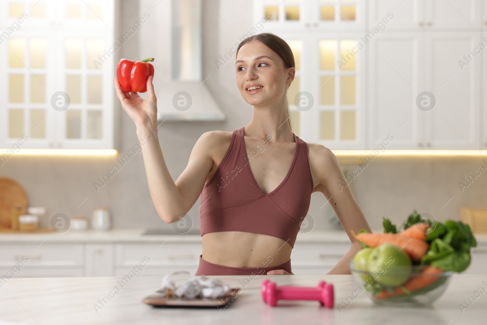 Woman holding bell pepper near table with kitchen scale, measuring tape, dumbbells and healthy food indoors Photo of Woman holding bell pepper near table with kitchen scale, measuring tape, dumbbells and healthy food indoors
