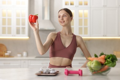 Photo of Woman holding bell pepper near table with kitchen scale, measuring tape, dumbbells and healthy food indoors