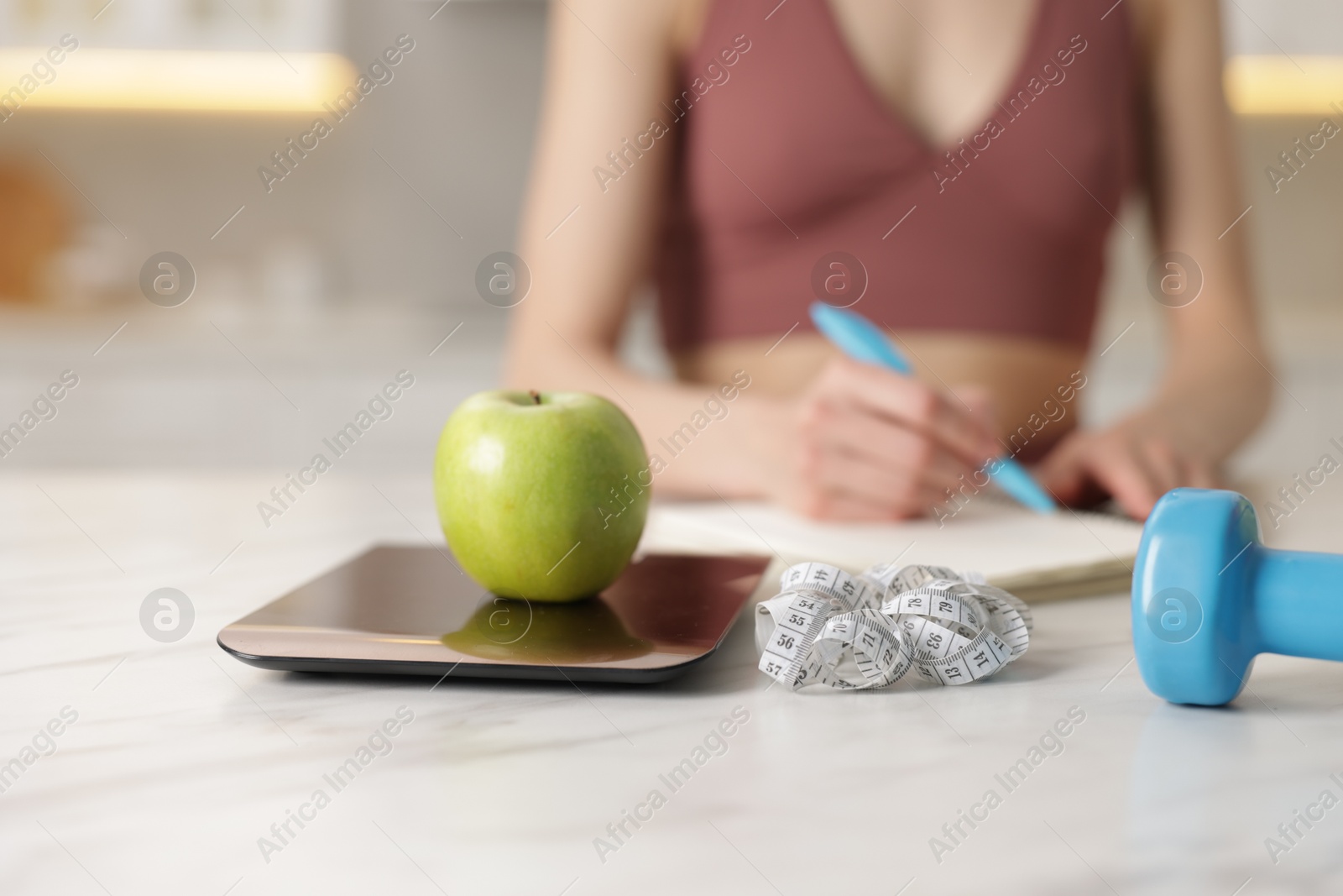 Photo of Woman developing diet plan at white marble table indoors, focus on kitchen scale, apple, measuring tape and dumbbell