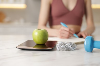 Photo of Woman developing diet plan at white marble table indoors, focus on kitchen scale, apple, measuring tape and dumbbell