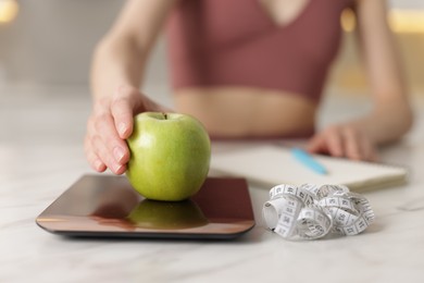Photo of Woman weighting apple on kitchen scale at white marble table indoors, closeup