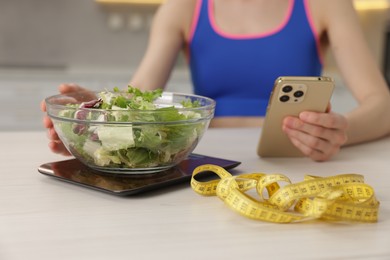 Photo of Woman with smartphone weighting salad on kitchen scale at white table indoors, closeup