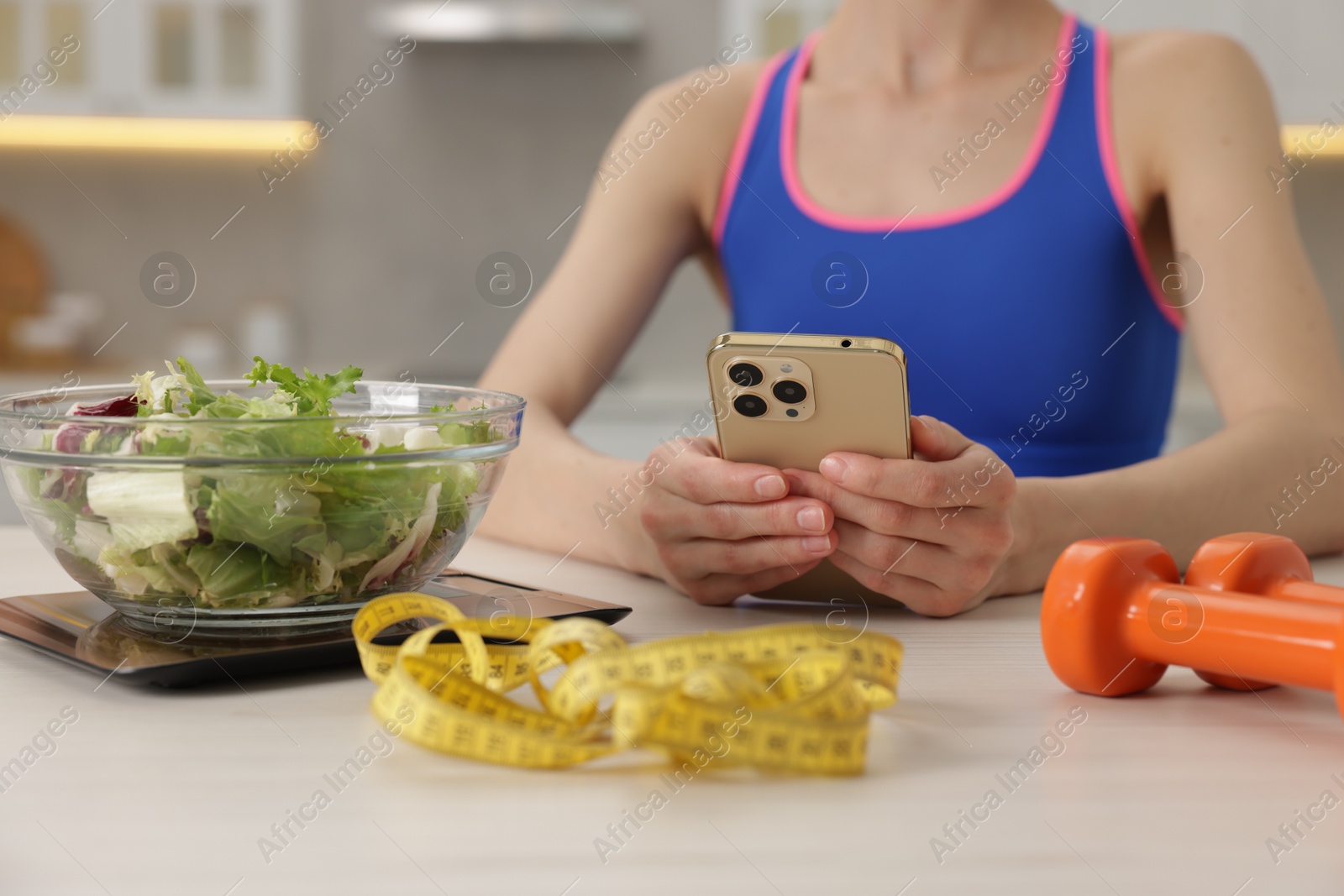 Photo of Woman using smartphone at white table with kitchen scale, salad, measuring tape and dumbbells indoors, closeup